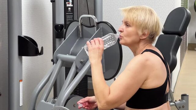 A woman takes a break to drink water while using gym equipment in the afternoon. She focuses on staying hydrated between exercises.