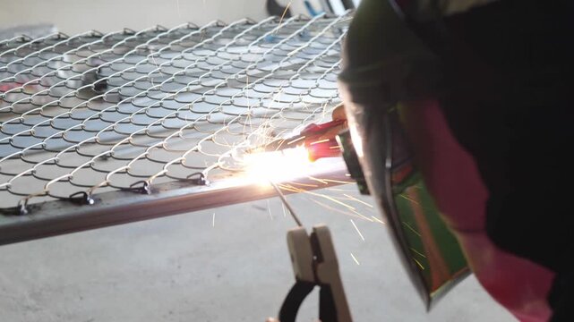 Welder wearing a protective face shield and gloves welds a steel rebar grid on the ground, with sparks and workshop tools visible in the background. Represents construction, metalwork, and industrial 