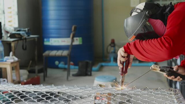 Welder wearing a protective face shield and gloves welds a steel rebar grid on the ground, with sparks and workshop tools visible in the background. Represents construction, metalwork, and industrial 