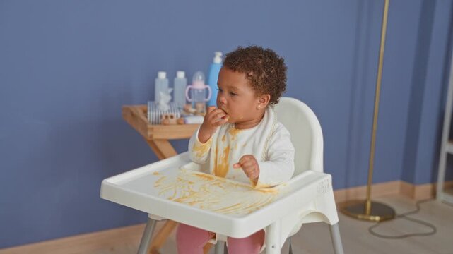 African american toddler girl in highchair eating with messy hands and tongue out in kitchen; playful curiosity.