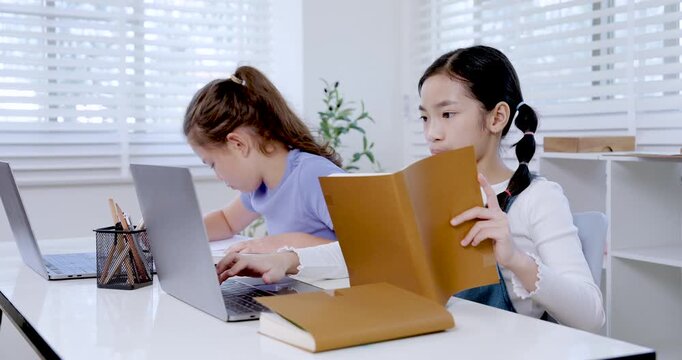 Asian preteen girl reading folder while caucasian girl typing on laptop beside her in classroom representing teamwork digital education learning project collaboration productivity childhood study
