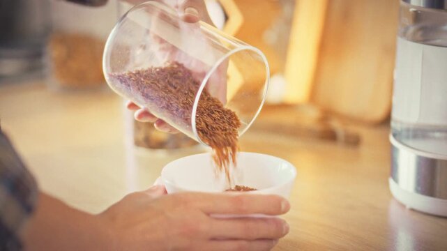 A housewife in a bright kitchen gently tips brown rice from a storage jar into a white bowl, beginning the simple ritual of measuring grains for cooking, highlighting everyday meal prep