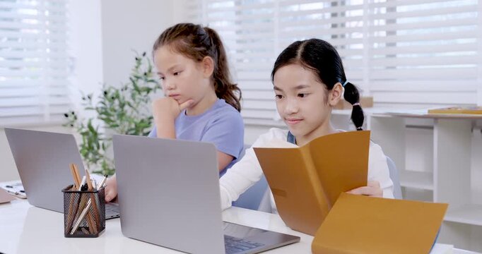 Asian preteen girl reading folder while caucasian girl working on laptop beside her in classroom representing teamwork digital learning collaboration education study productivity project communication