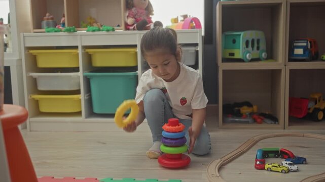 Girl stacking plastic rings toy while kneeling and reaching with hand in playroom; concentration learning play.
