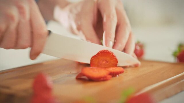 A man slices ripe red strawberries into thin slices on a cutting board in a home kitchen, preparing the sweet, juicy berries for dessert, yogurt, or smoothies. Healthy eating concept
