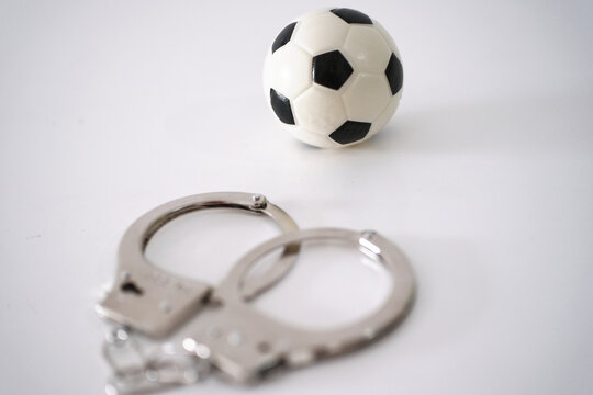 A miniature soccer ball resting on a pair of metal handcuffs, conceptualizing sports crime, match-fixing, corruption, or legal issues in football.