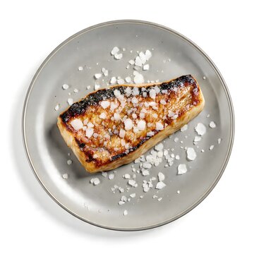 Perfectly Seared Salmon Fillet: A succulent salmon fillet, expertly seared to a golden-brown crisp, sits atop a minimalist grey plate, generously seasoned with coarse sea salt flakes.