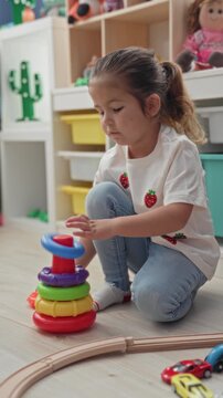 Young hispanic girl seated on floor playing, stacking a multicolored plastic ring toy and placing rings with hand in a playroom building; playful learning.