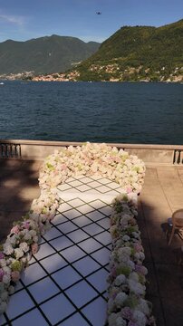 Luxury outdoor wedding ceremony setup on Lake Como, Italy. White aisle leading to a floral arch with fresh blooms, surrounded by guest seating and panoramic lake and mountain views