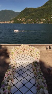Luxury outdoor wedding ceremony setup on Lake Como, Italy. White aisle leading to a floral arch with fresh blooms, surrounded by guest seating and panoramic lake and mountain views