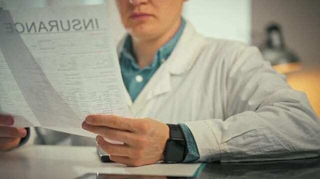 Examining patient documents, doctor with glasses works in bright medical office, consulting patient card and insurance forms while meticulously reviewing records, showcasing careful clinical analysis