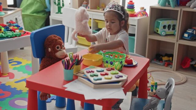 Hispanic girl wearing a tiara plays tea party with a stuffed teddy, lifting a plush while seated at a small play table in a building; playful imagination joy.
