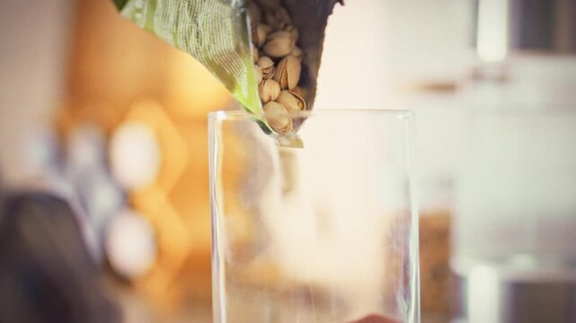 In a bright home kitchen, a housewife carefully transfers nuts and salted pistachios from a grocery bag into a clear glass jar, organizing her snack supply and emphasizing healthy food habits