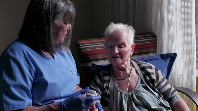 Female nurse using a pulse oximeter on an elderly woman's finger during a home visit, providing compassionate medical care and health monitoring for the senior patient sitting in a chair