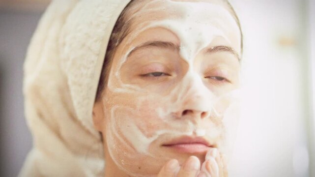 In bathroom, young girl wrapped in cozy robe stands before mirror, applying white cleansing foam to her face with gentle strokes, a serene moment in her daily beauty routine, aided by a nearby towel