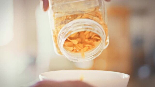 In bright home kitchen, young woman's hands scoop yellow cornflakes into a white bowl, preparing a low-calorie breakfast. The mindful ritual highlights conscious nutrition and figure-friendly eating