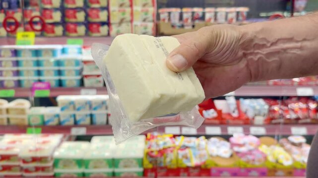 Customer's Hand Inspecting a Cheese Package While Shopping in a Grocery Store Aisle.Close-Up of a Man's Hand Spinning a Pack of Cheese While Browsing Products in a Supermarket Refrigerator Section