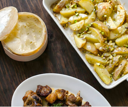 Patatas asadas con pistachos servidas con queso fundido tipo torta y carne cocinada sobre mesa de madera