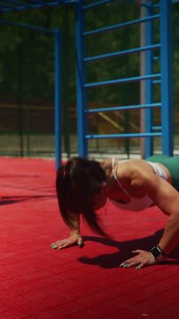 A fit woman performs push-ups on a vibrant red outdoor workout mat. She is focused on her physical training in a sunny outdoor gym.