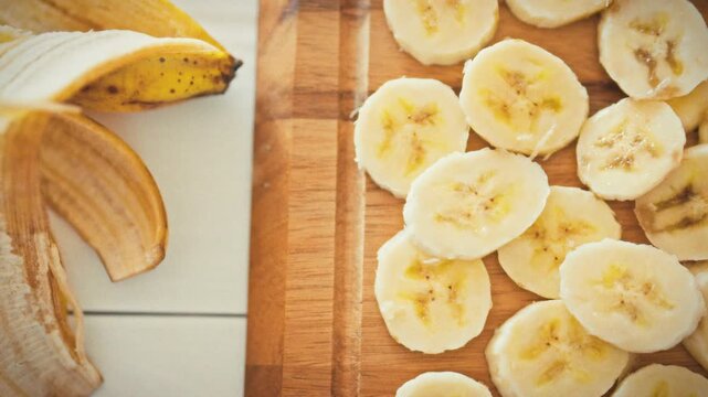 On a white wooden table, a banana peel lies next to sliced yellow banana rounds on a wooden cutting board, highlighting a food scene that emphasizes nutrition, health and balanced eating