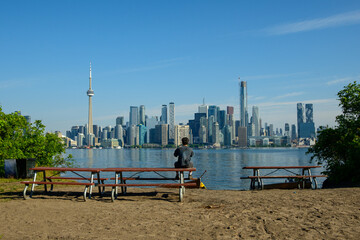 Obraz premium Person sitting on a picnic table photographing the Toronto skyline from a sandy island shore.