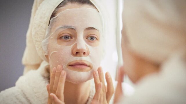 To achieve radiant glow, a girl in a white robe with a towel on her head stands before the bathroom mirror, smoothing an aloe-vera sheet mask over her face to lock in moisture and pamper skin at home
