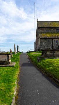 An ancient church made of dark stone, with a moss-covered roof and a surrounding cemetery. A path separates the cemetery and the church