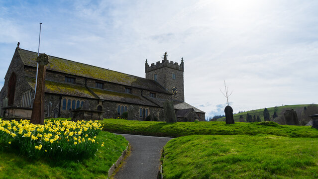 St Michael & All Angels Church, Hawkshead, Lake District, England. A stone church with a distinctive crenellated tower and a clock on it surrounded by green grass and paths