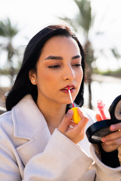 A young woman applies lip gloss on a city street during a sunny day
