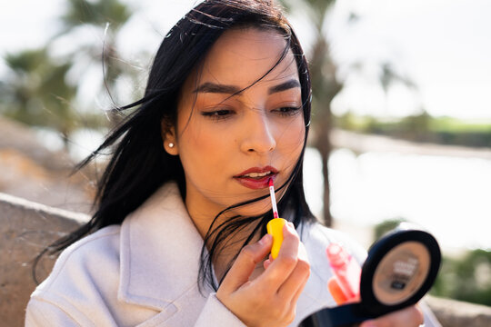 A young hispanic woman is applying lip gloss on a city street