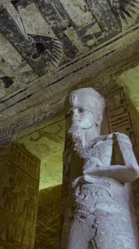 Grand hypostyle hall of Abu Simbel temple, Egypt, with two rows of colossal Ramesses II osiride figures flanking painted ceiling covered in vultures, cartouches and hieroglyphics.