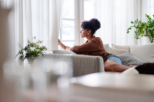 Woman gazes out window from couch in bright living room.
