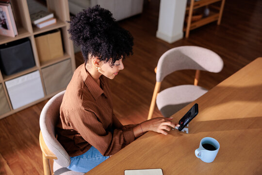 Woman on smartphone video call with coffee at home dining table.