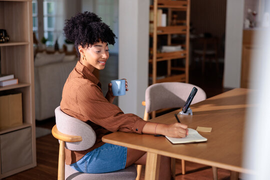 Woman taking notes with coffee while working from home on smartphone