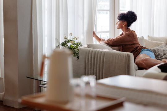 Woman looking through window curtains while resting on sofa at home
