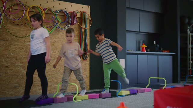 Young children are actively participating in an agility and fitness training session. Kids practice coordination and balance using hurdles and blocks in a gym.