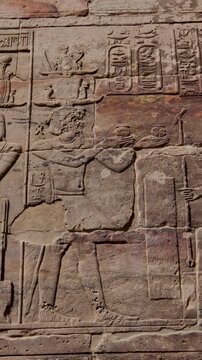 Exterior sandstone wall of Philae temple, Aswan, Egypt, with large carved relief depicting seated gods and pharaoh offering ritual ceremony, hieroglyphic cartouches under vivid blue sky.