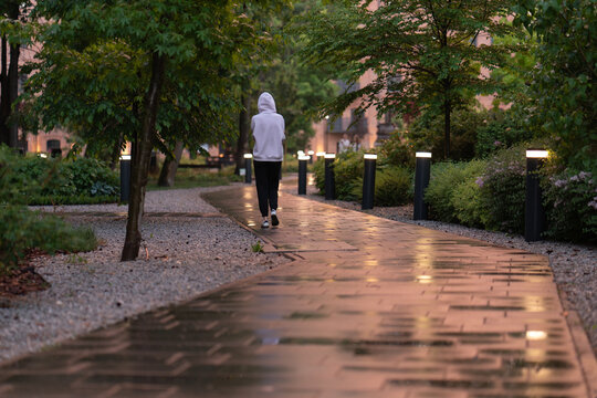 A lonely person in a white hoodie walks along a wet cobblestone path at dusk, surrounded by modern outdoor lighting, green trees, and shrubs in a peaceful residential courtyard or urban park.