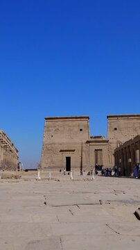 Tourists exploring the Philae Temple complex under clear blue sky, showcasing ancient Egyptian architecture, courtyards, and well-preserved stone structures.