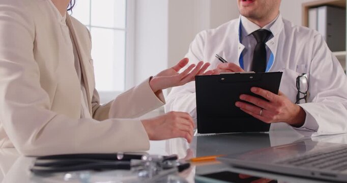 Unknown doctor giving consultancy to woman patient, filling out medical history form, listening to complaints, writing down prescriptions, sitting at desktop with sthetoscope and laptop computer