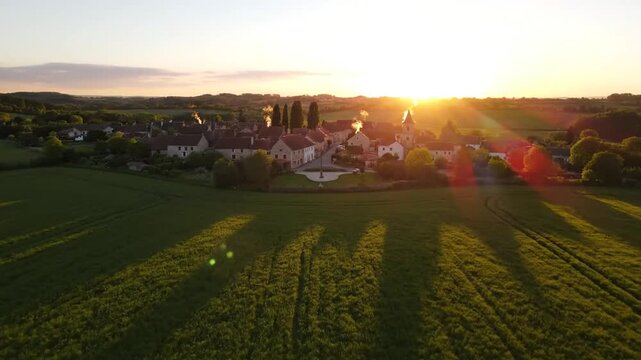 Golden hour aerial view of a quaint European village nestled among rolling green fields with a church steeple and soft sunset light casting long shadows