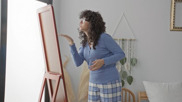 Woman adjusting hair at a freestanding mirror in a cozy bedroom, wearing pajama top and plaid lounge pants while touching curls; calm private routine.