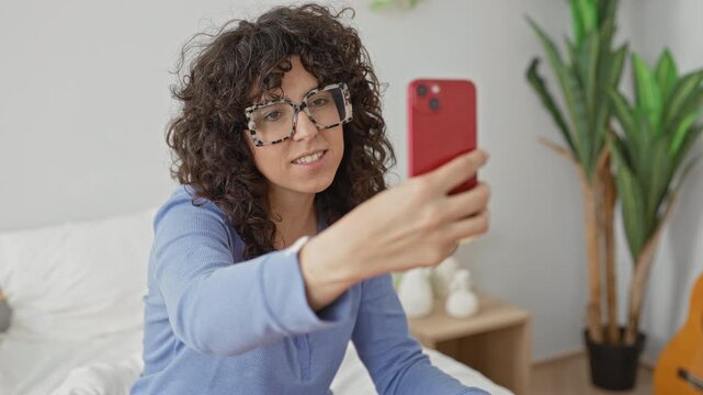 Woman holds red smartphone with outstretched arm taking a selfie on bed in building; confidence candid selfcare.