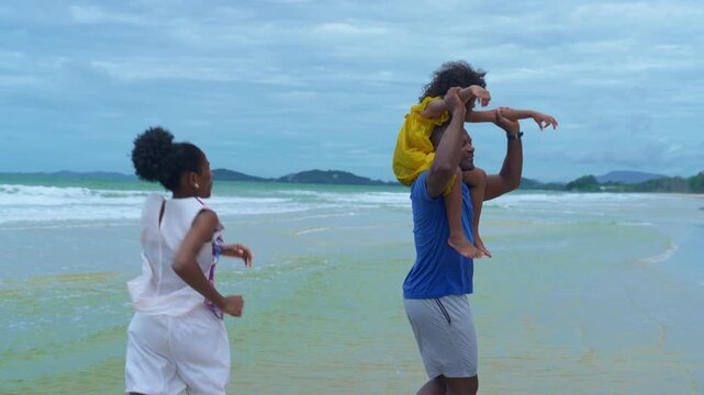 Beach family joyful scene with black hair father mother child at ocean, wave rolling under cloudy sky, shoulder carry while mother runs nearby, child walking shoreline, carefree warm bond