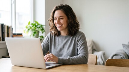Fototapeta premium Smiling young woman with wavy brown hair working on laptop at desk in cozy room. Happy female freelancer or student studying online, using wireless earphones and drinking coffee.