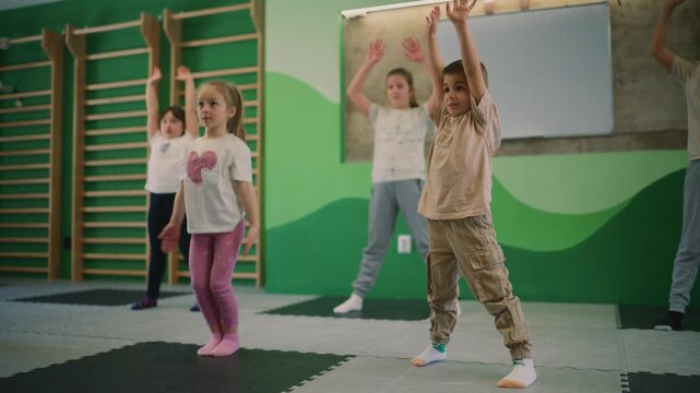 Young children are performing jumping jacks during a fitness class indoors. Kids are actively engaging in physical education exercises for their health.