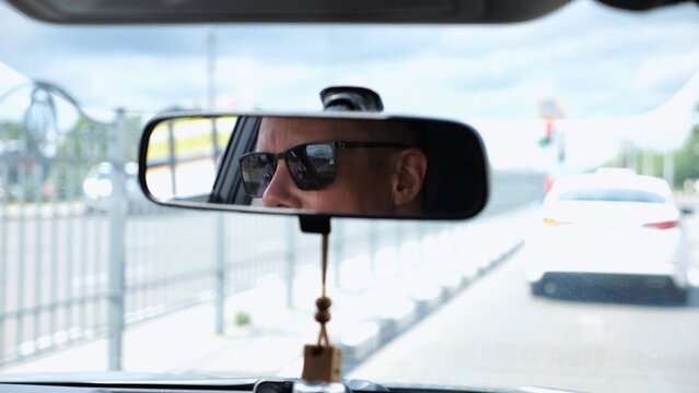 Eyes in sunglasses of male driver reflecting in rearview mirror while riding motor auto. Young guy looking concentrated to urban road during operating vehicle. Confident man driving modern car at city