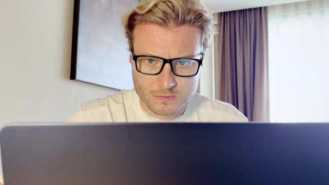 Close-Up of a Young Man Working on a Laptop, Wearing Glasses with the Screen Light Reflecting in His Lenses