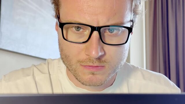 Close-Up of a Young Man Working on a Laptop, Wearing Glasses with the Screen Light Reflecting in His Lenses