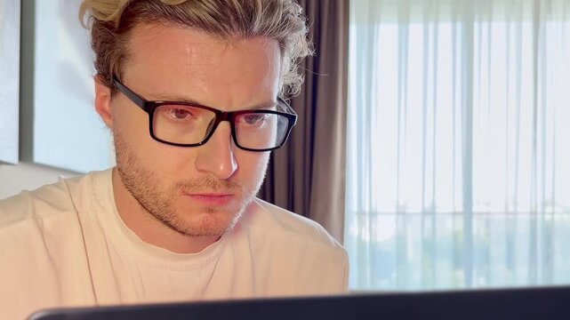 Close-Up of a Young Man Working on a Laptop, Wearing Glasses with the Screen Light Reflecting in His Lenses